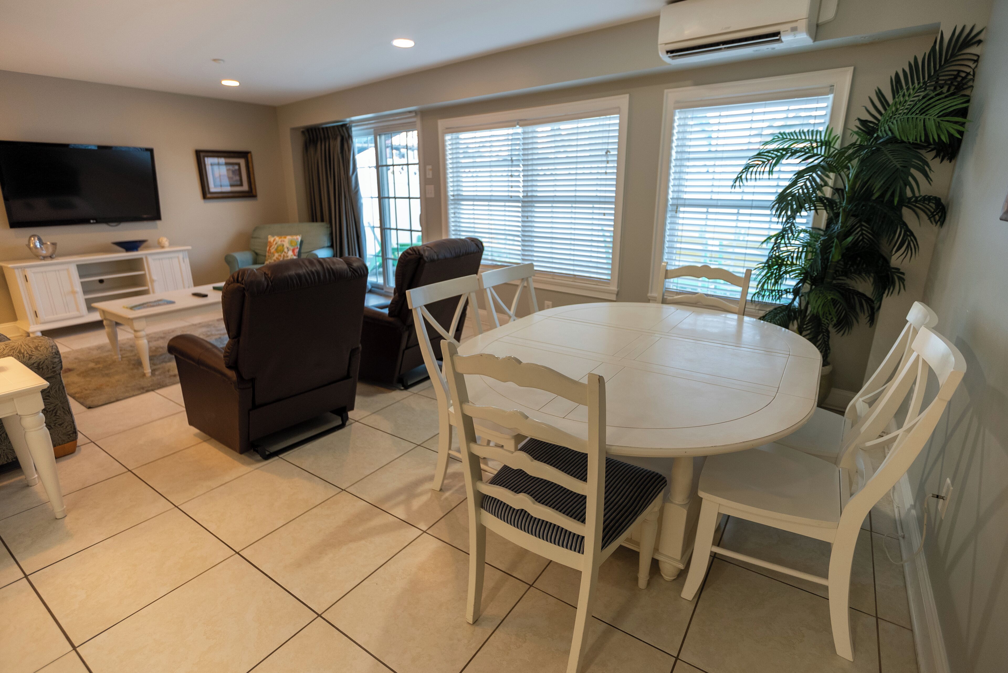 A living room with a white round table, white chairs, a brown sofa, a TV, and a large window with blinds.