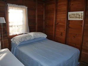 Une pièce de style cabane en bois, avec un lit, une fenêtre ornée d’un rideau blanc et une photographie encadrée accrochée au mur.