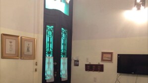 Quarto com porta preta, um candelabro, televisão fixada na parede e uma pequena mesa com uma cadeira.