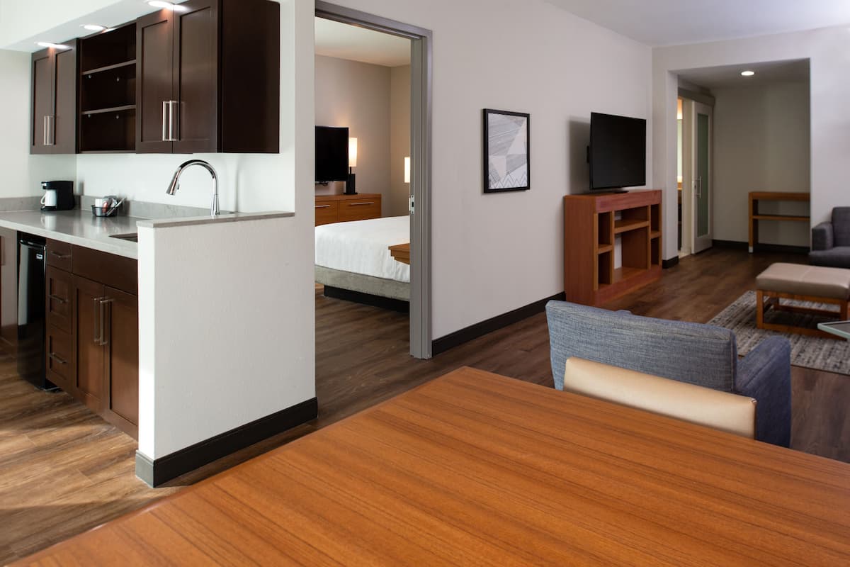 A modern hotel room with a kitchenette, a dining area, a living room with a sofa and TV, and a bedroom with a bed and nightstand.