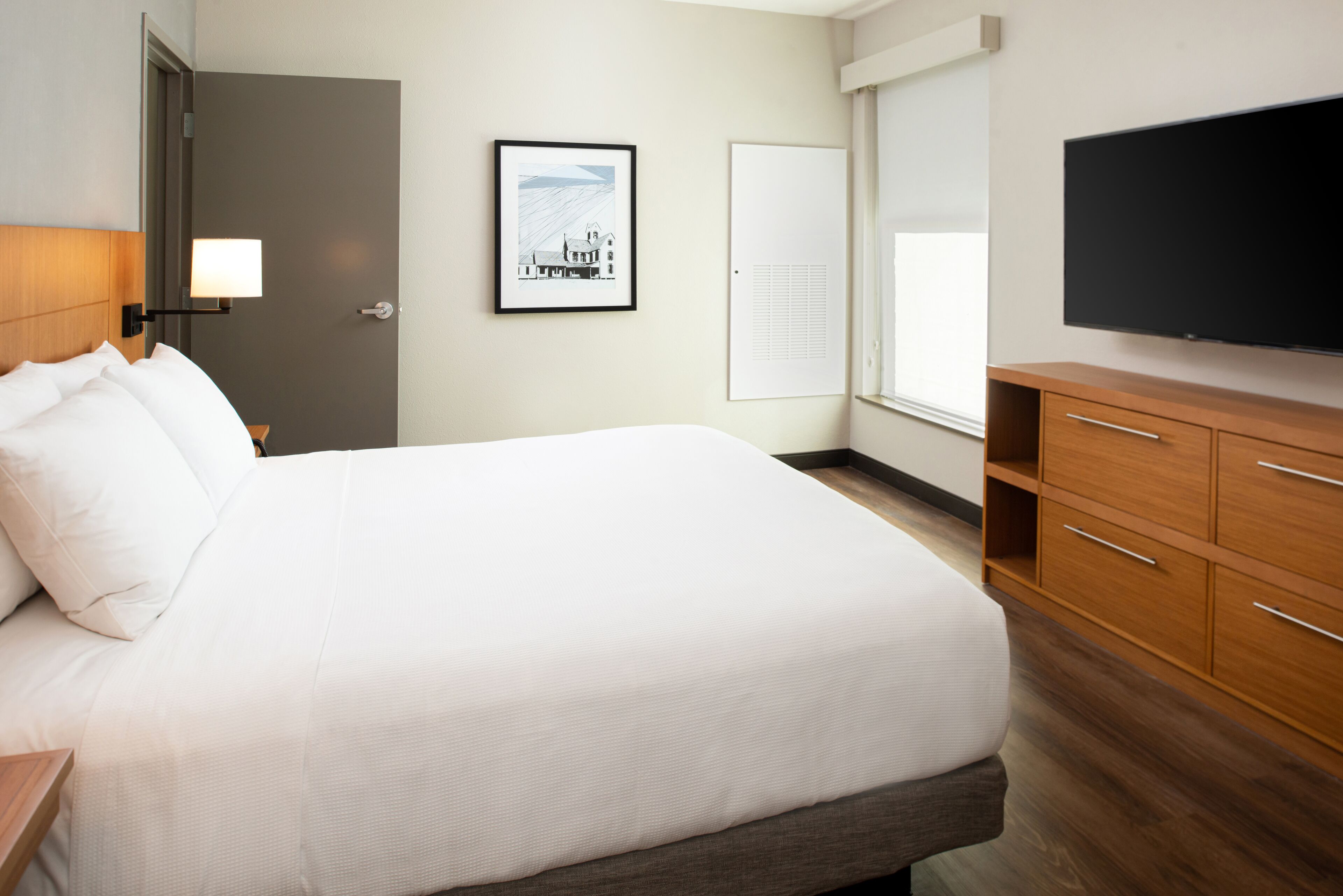 Ein Hotelzimmer mit einem großen Bett, Nachttischen aus Holz, einem Flachbildfernseher, einem gerahmten Bild an der Wand und einem Fenster mit Jalousien.
