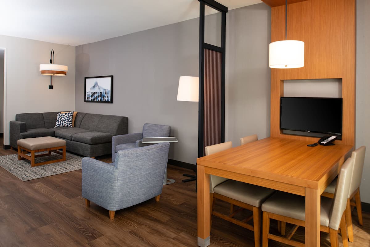 A modern living room with a grey sofa, a blue armchair, a wooden dining table, and a flat-screen TV mounted on the wall.