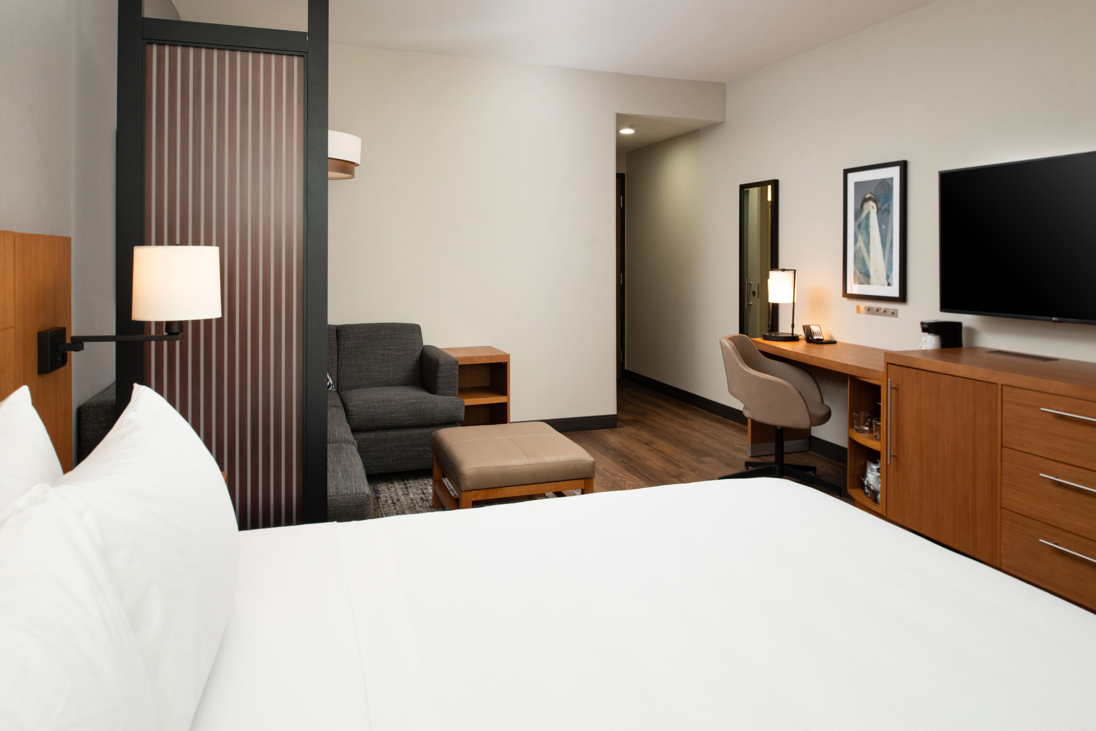 Ein Hotelzimmer mit Bett, Schreibtisch, Sessel, kleinem Hocker, Lampe, Fernseher und einem gerahmten Bild an der Wand.