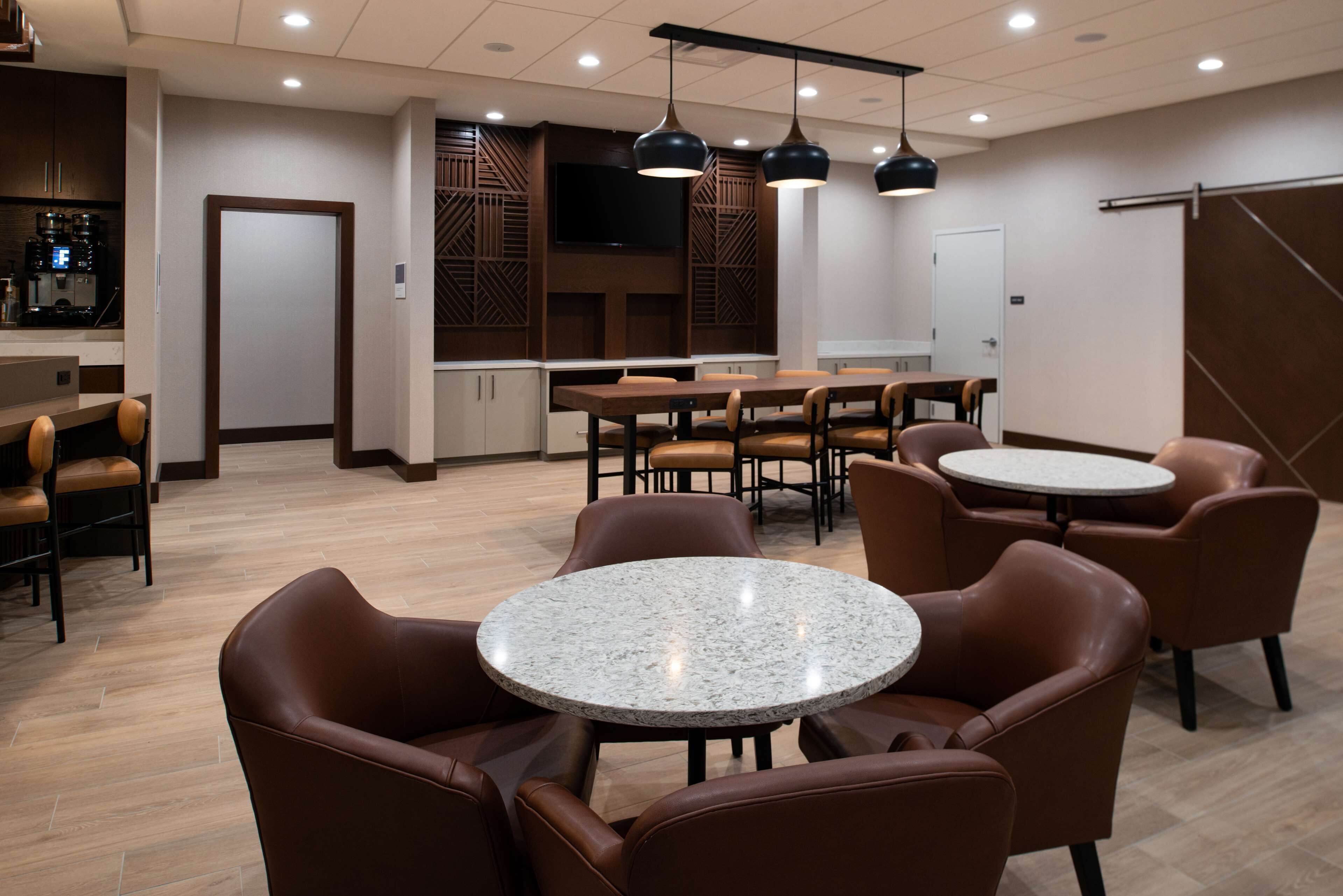 Un comedor moderno con mesas redondas y sillas de cuero marrón, una zona de bar con una máquina de café y un televisor de pantalla plana grande en la pared.