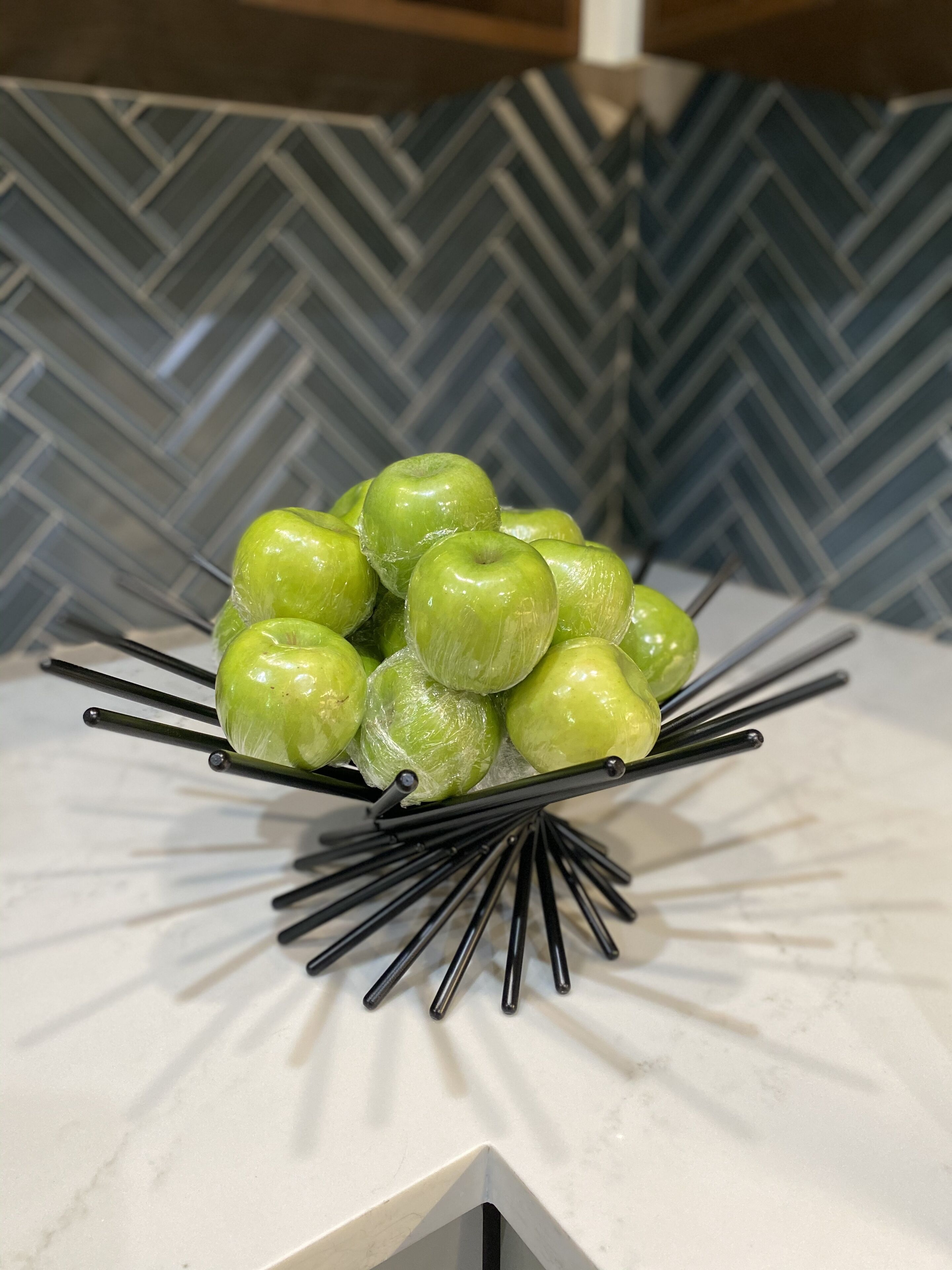 Un bol de pomes verdes sobre una taula de marbre blanc amb un fons de rajoles de disseny geomètric.
