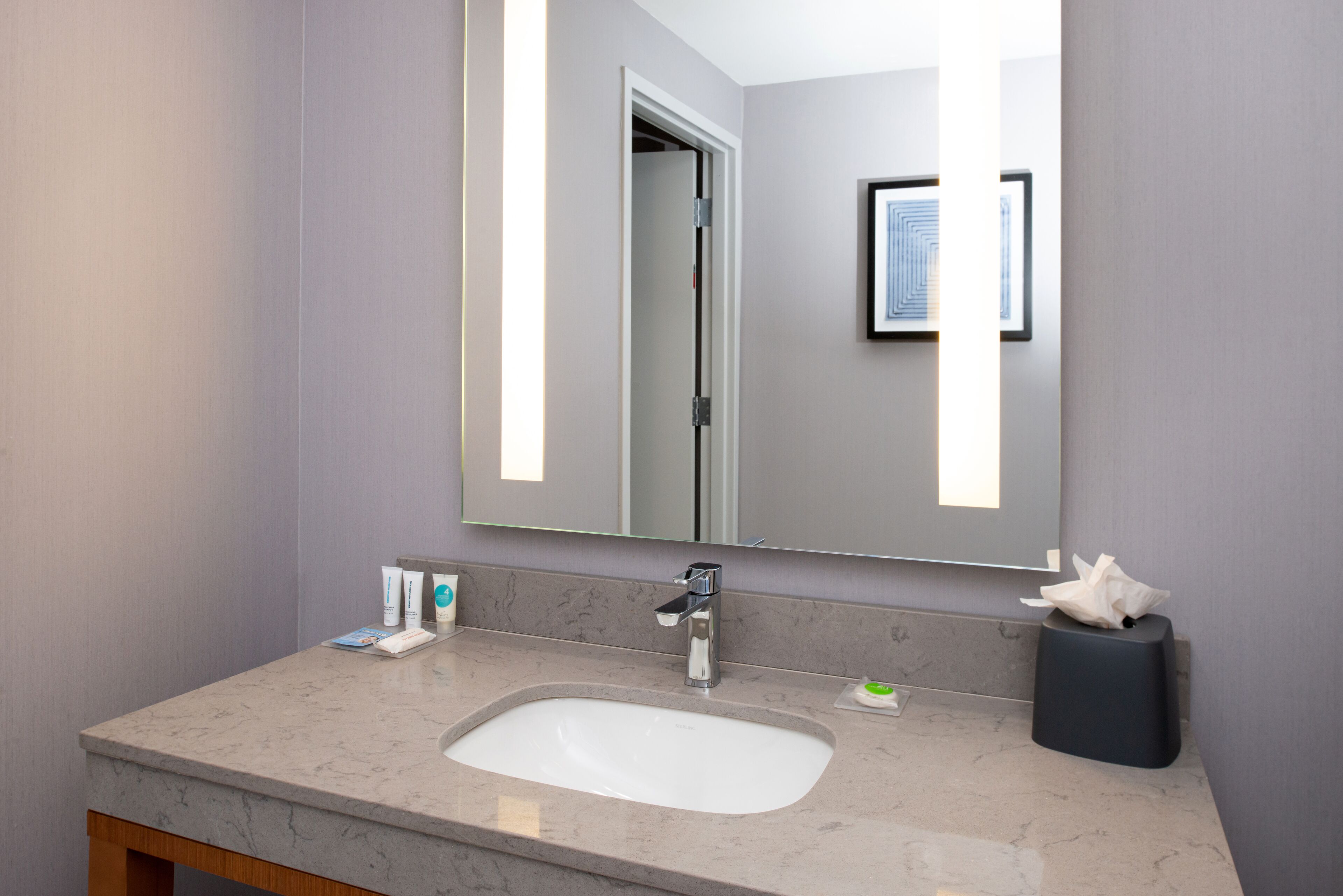 A modern bathroom with a marble sink, a large mirror, and a framed artwork on the wall.