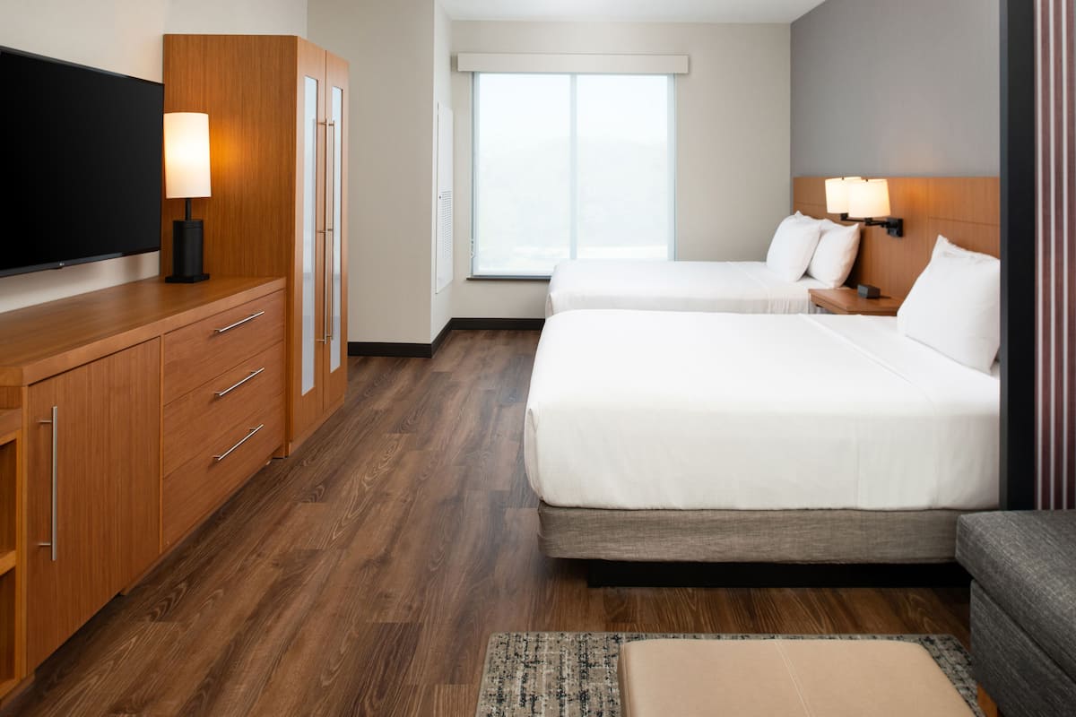 A hotel room with two beds, a TV, wooden furniture, a window with blinds, and a nightstand with lamps.