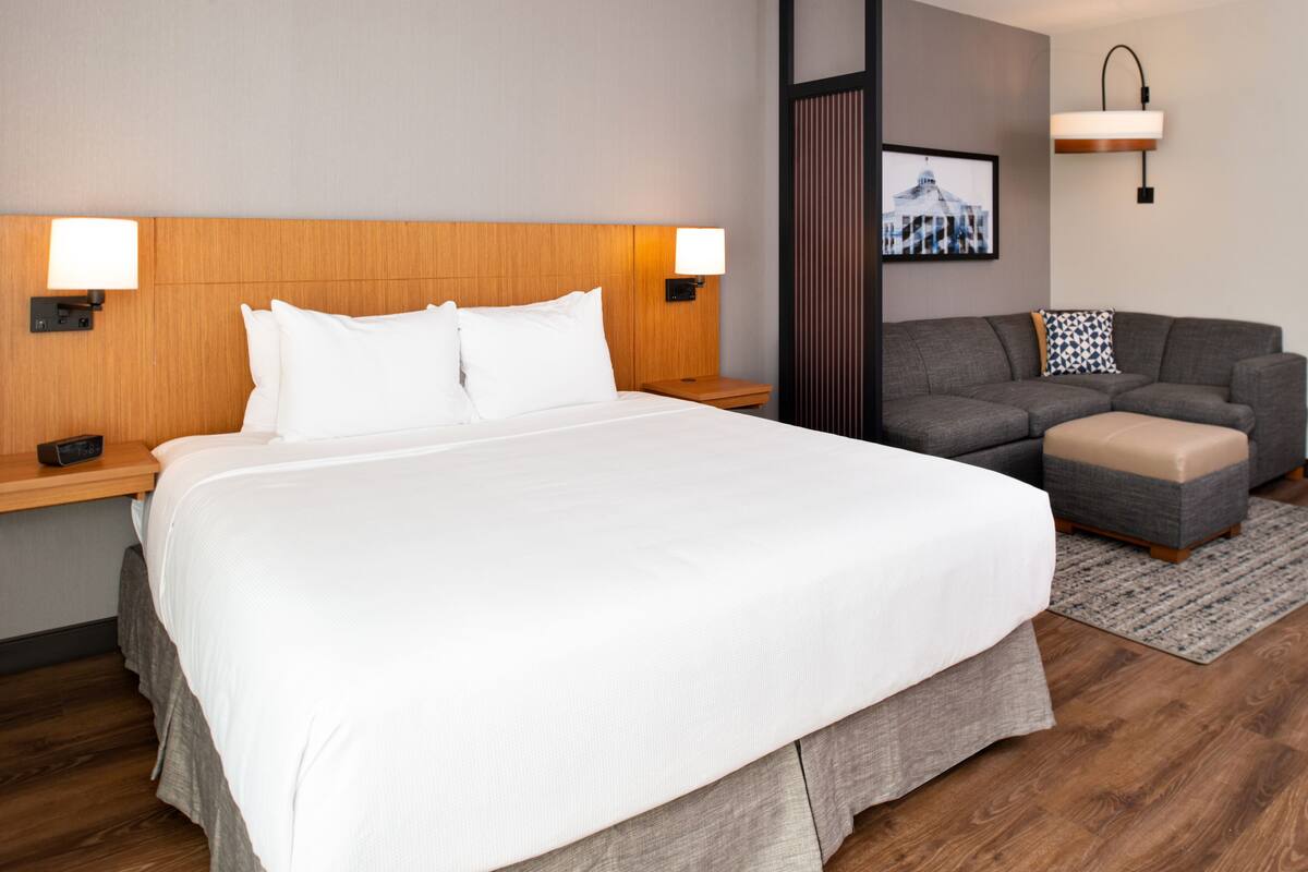 A hotel room with a large bed, a grey sofa, a small ottoman, and two bedside lamps.