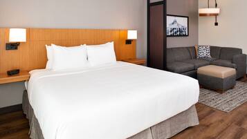 Una habitación de hotel con una cama grande, un sofá gris, un pequeño otomaní y dos lámparas de noche.