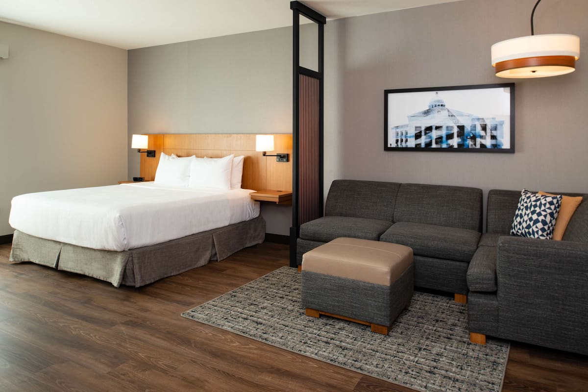 A hotel room with a large bed, a sofa, a small ottoman, a nightstand, a lamp, and a framed picture on the wall.