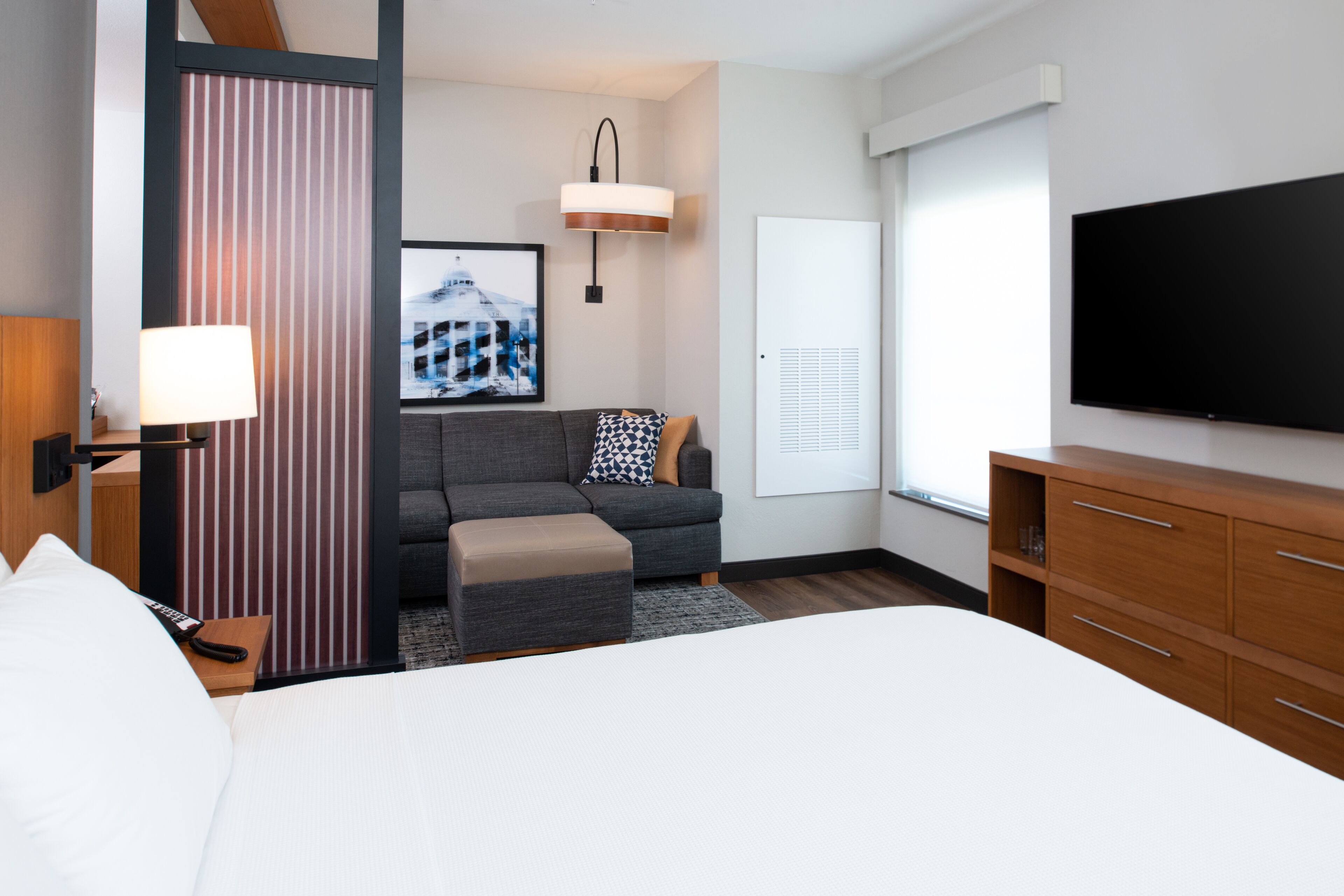 Quarto de hotel moderno com uma cama grande, um sofá cinzento, uma cômoda de madeira, uma TV de tela plana, um quadro na parede e um abajur em uma mesa de cabeceira.