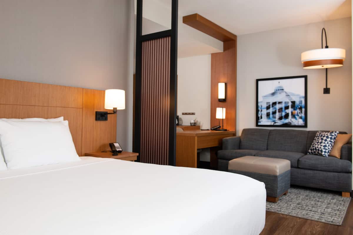 A hotel room with a large bed, a desk, a sofa, and a chair. There is a lamp on the desk and a picture on the wall.