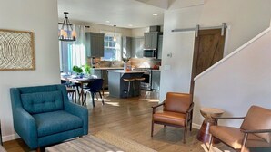 A modern living room with a sofa, coffee table, and chairs. A kitchen with a dining area is visible in the background. - Bluebird Day - River Trail House, Enjoy a cozy retreat in Bend, OR. (Bend)