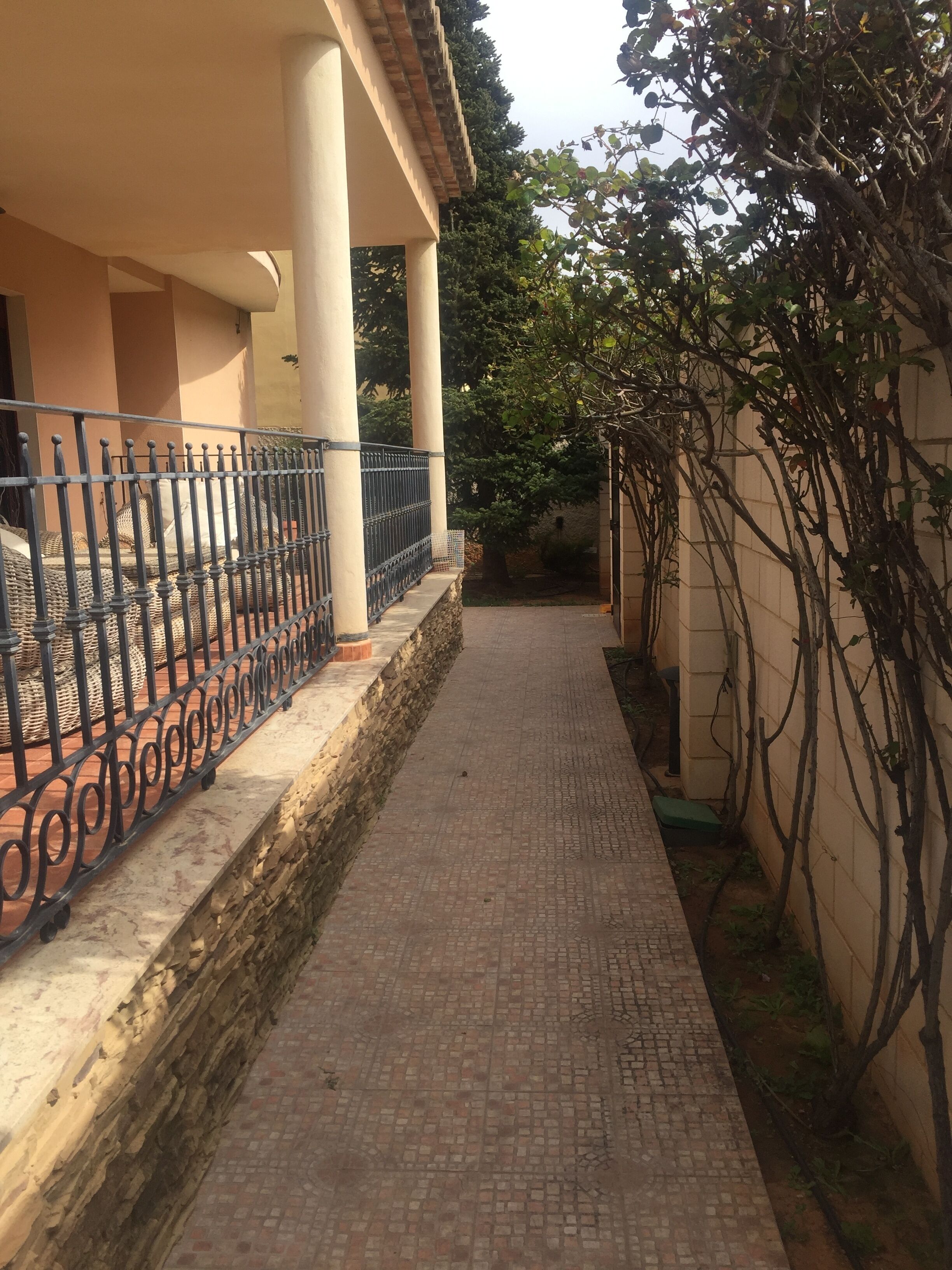 A covered walkway with a brick floor, bordered by a stone wall and a metal fence.
