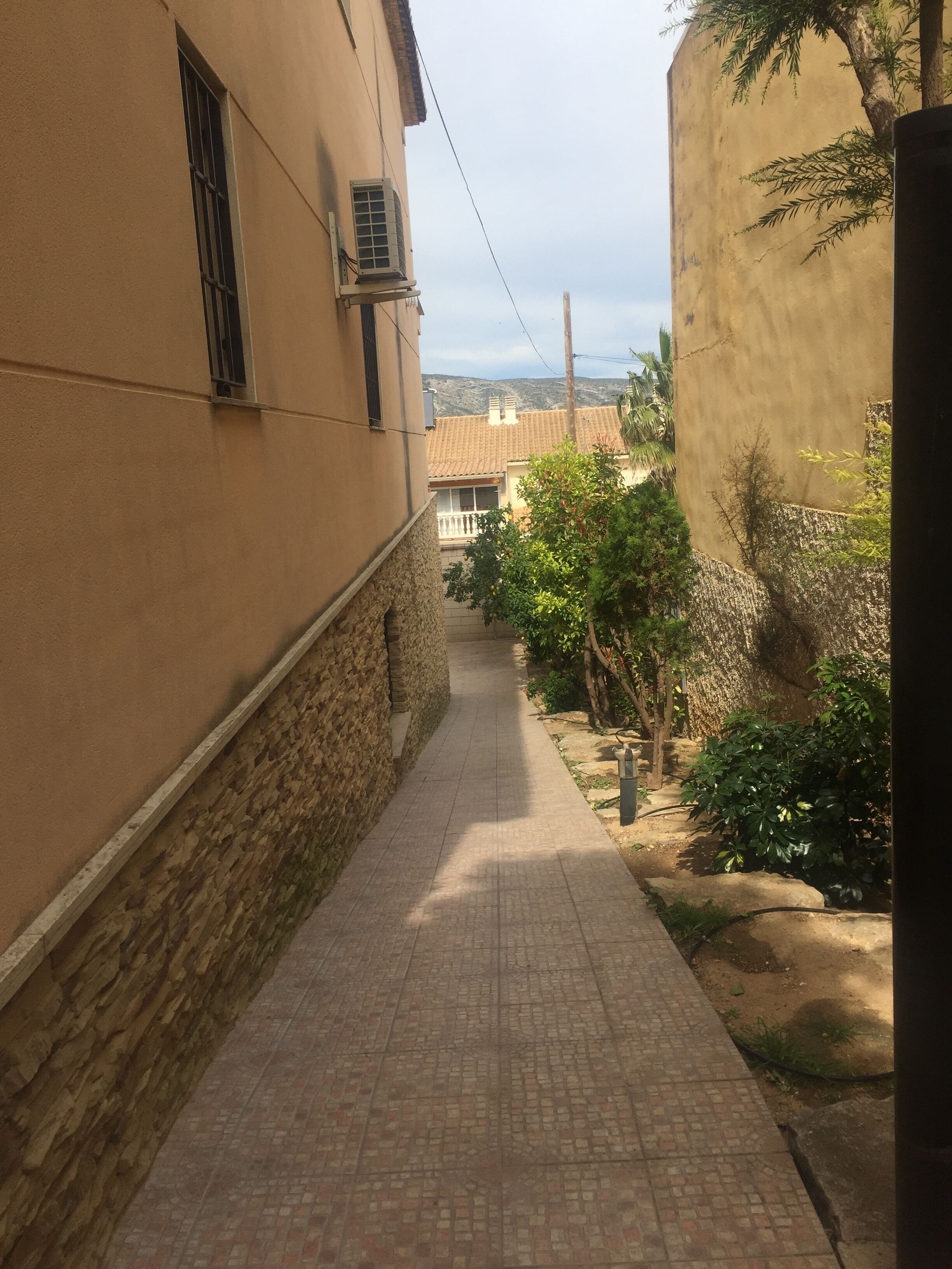 A narrow paved pathway flanked by buildings with stone and plaster walls, leading to a residential area with greenery and a distant view of hills.