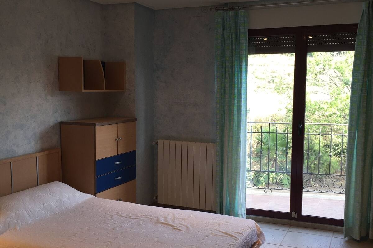 A bedroom with a bed, a wardrobe, a radiator, and a balcony with a view of greenery.