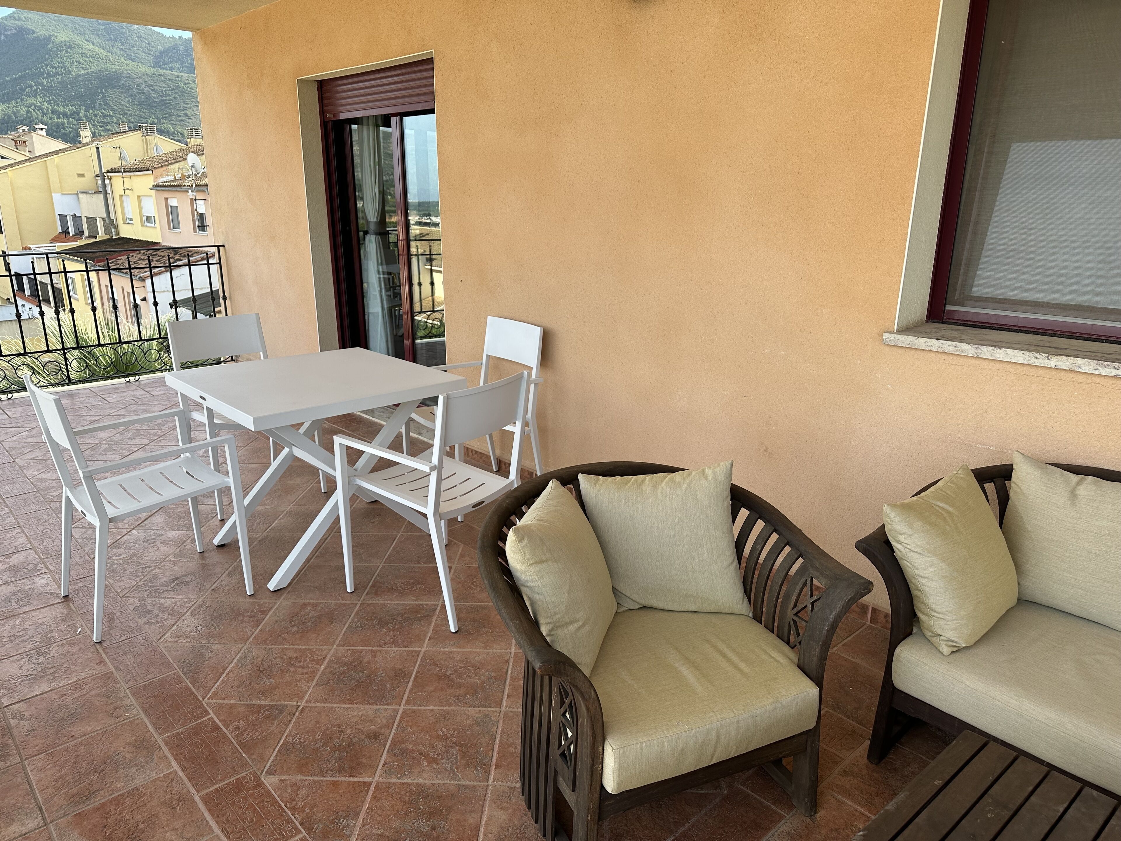 A balcony with a white table and chairs, a wicker sofa with cushions, and a view of buildings and hills in the background.