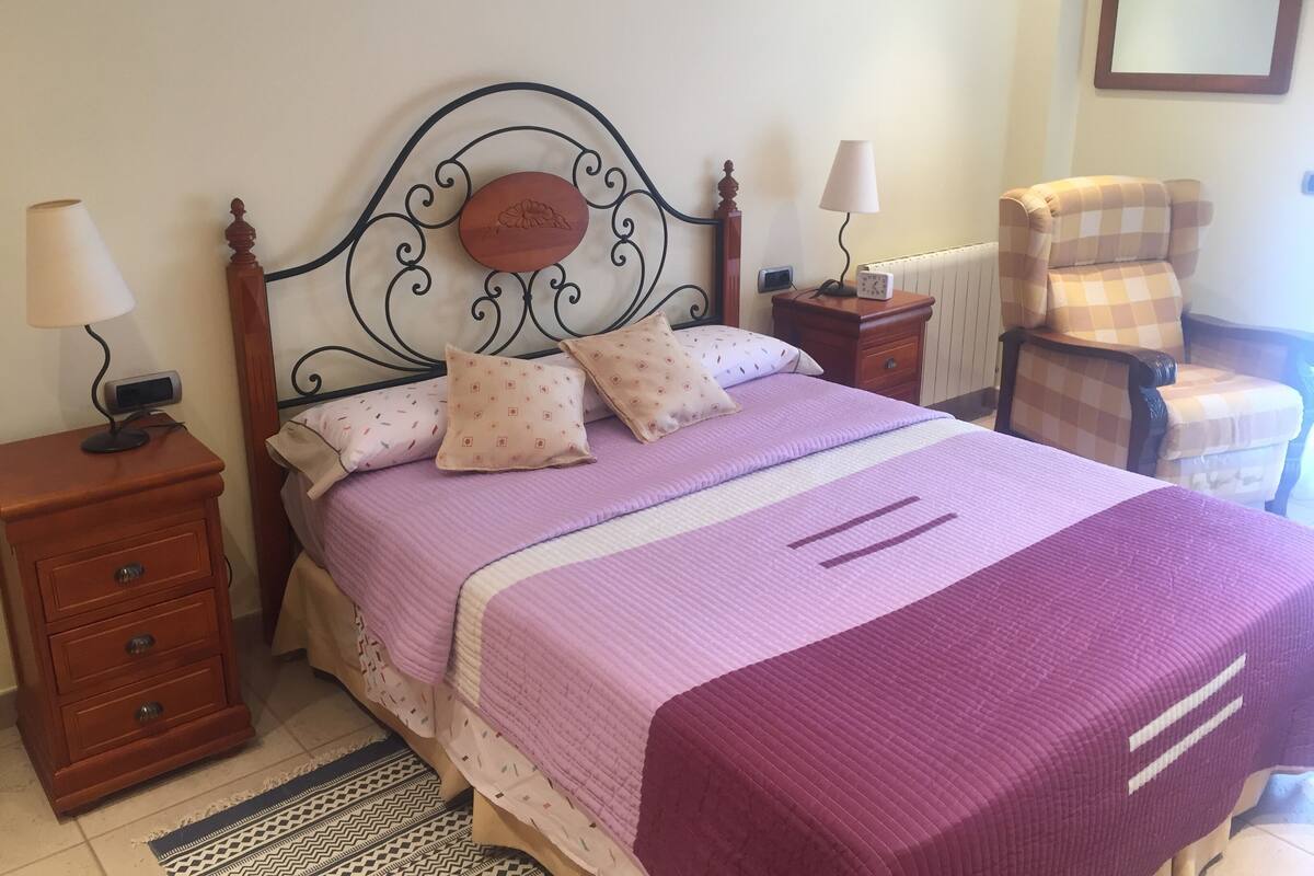 A neatly made bed with a purple and white bedspread, two patterned pillows, a bedside table with a lamp, and a chair with a checkered cushion.