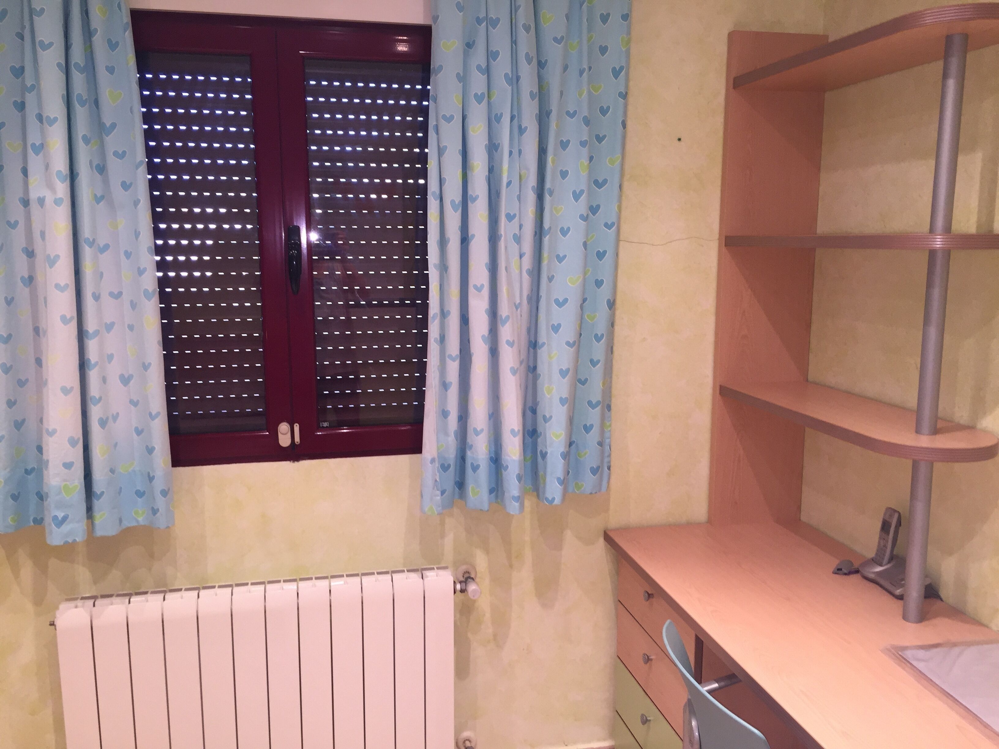 A bathroom with a window, a radiator, a wooden shelf, and a mirror.