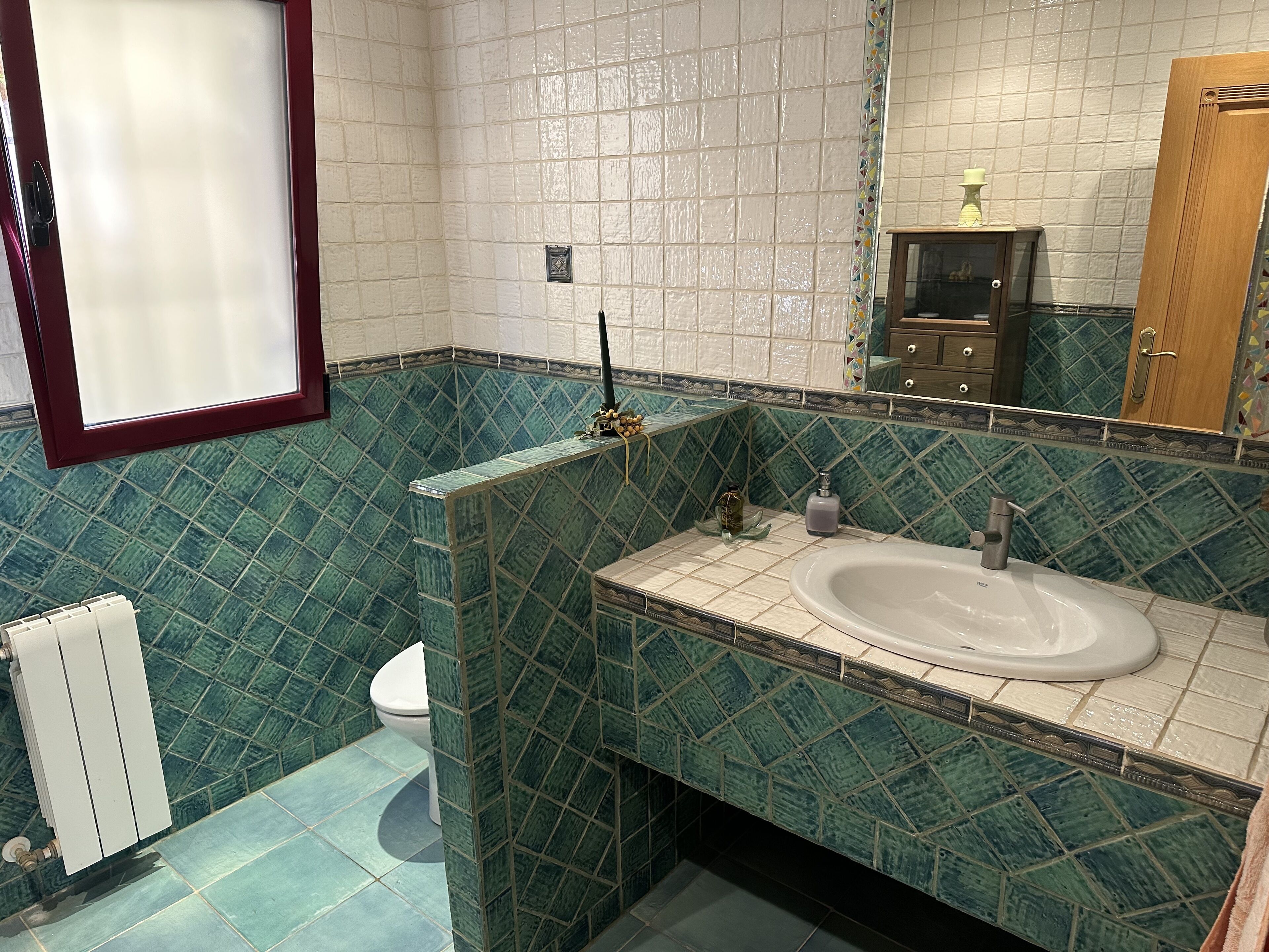 Une salle de bain avec un lavabo blanc, des carreaux verts à motifs, des toilettes et un miroir avec un cadre en bois.