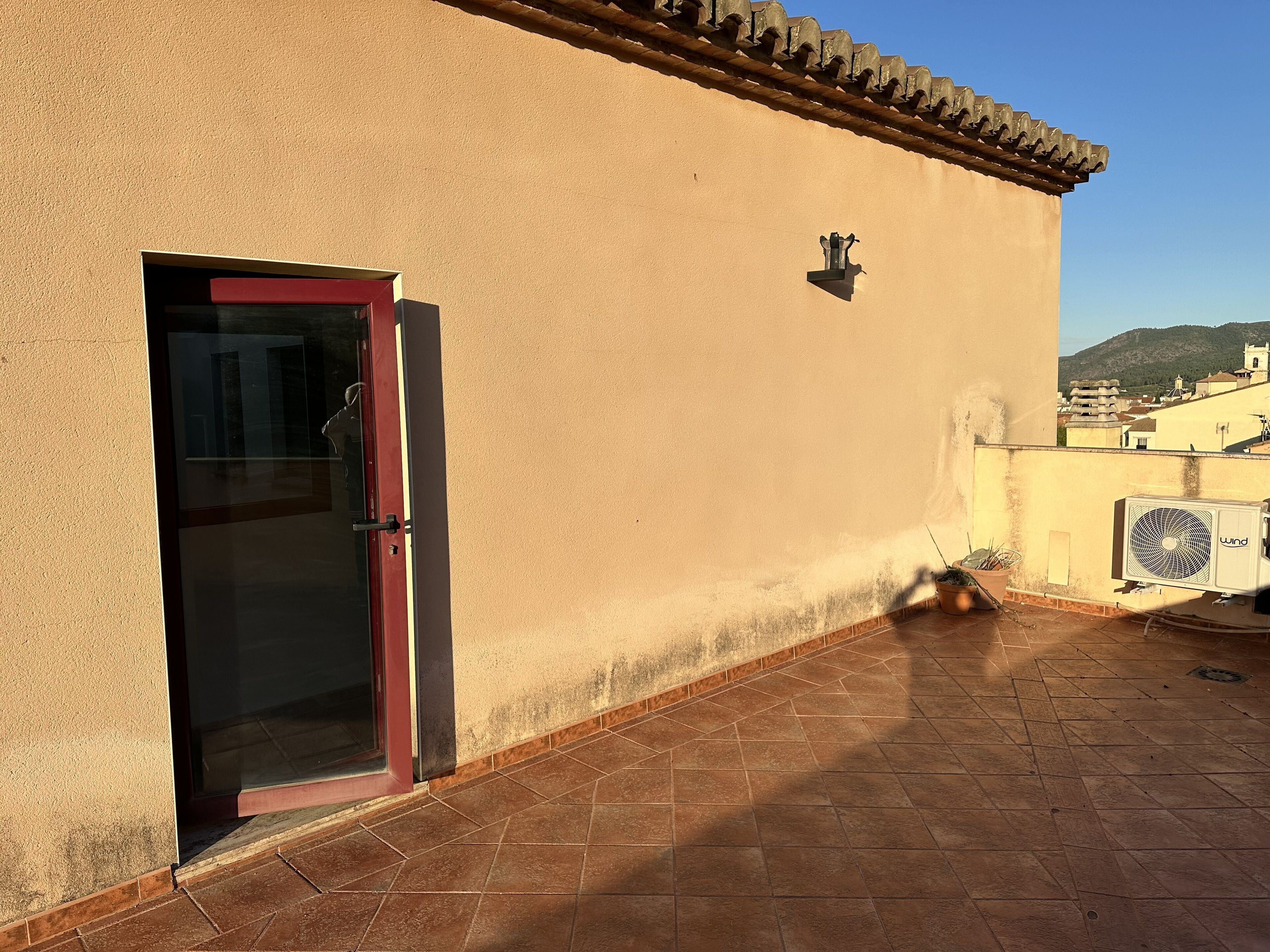 Un bâtiment avec une porte rouge, un petit espace extérieur agrémenté d’un pot de fleurs et un appareil de climatisation fonctionnant à l’énergie éolienne.