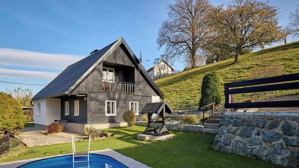 Une maison à deux étages, à l’extérieur sombre, avec un mur en pierre et une piscine en premier plan.