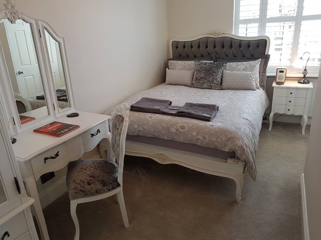 A bedroom with a bed, a vanity with a mirror, a chair, and a bedside table with a lamp.
