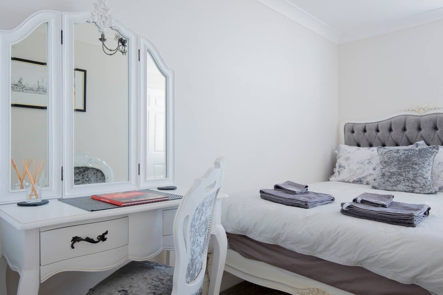 Un dormitorio con un tocador blanco, una cama con ropa de cama gris y blanca, y una silla con estructura blanca.