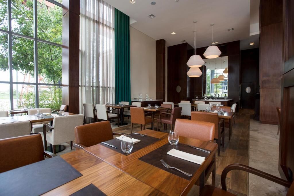 A modern restaurant with wooden tables and chairs, large windows with curtains, and pendant lighting.
