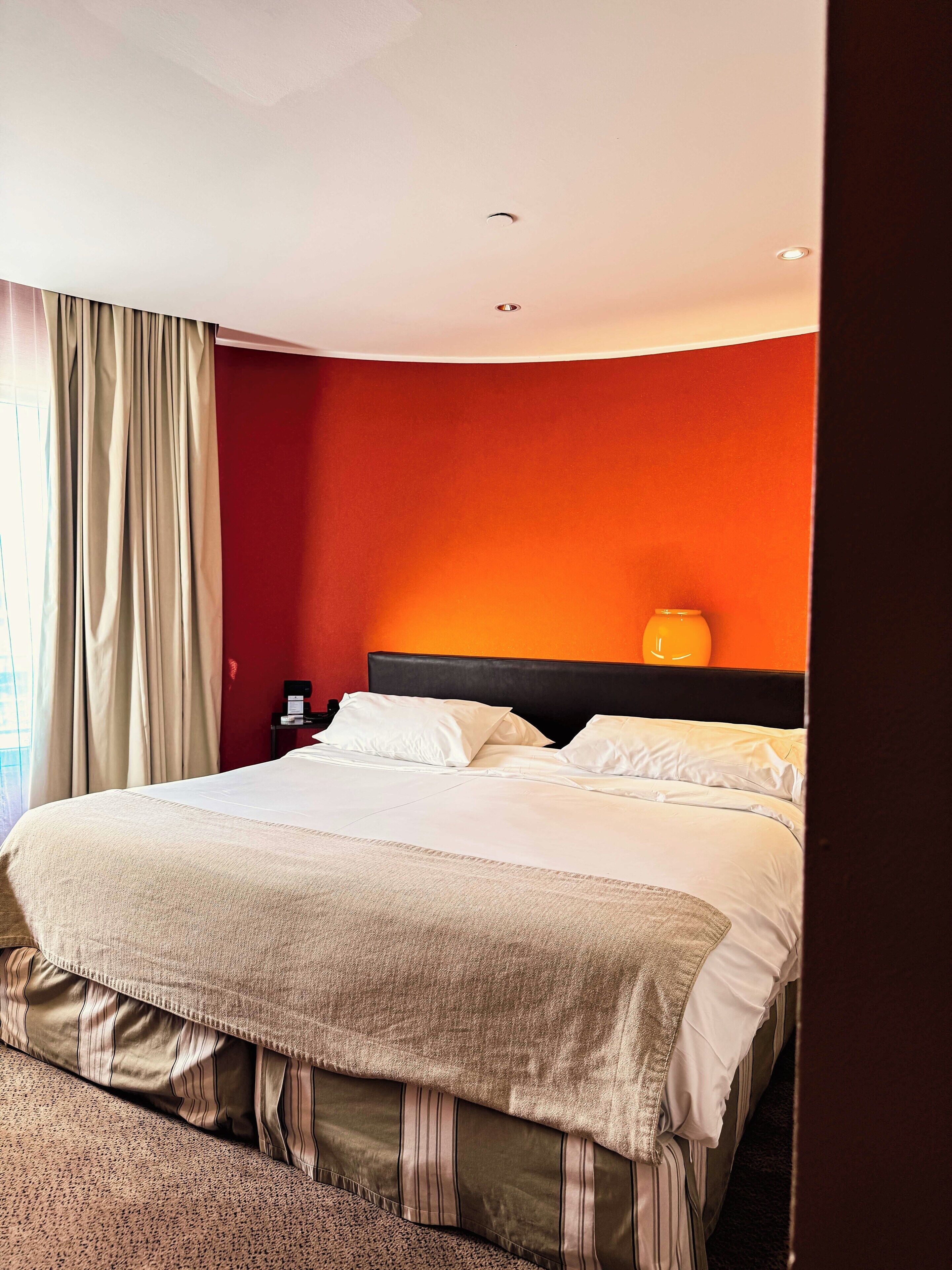 Una habitación de hotel con cama, cortinas, una mesita de noche y una lámpara de pared.