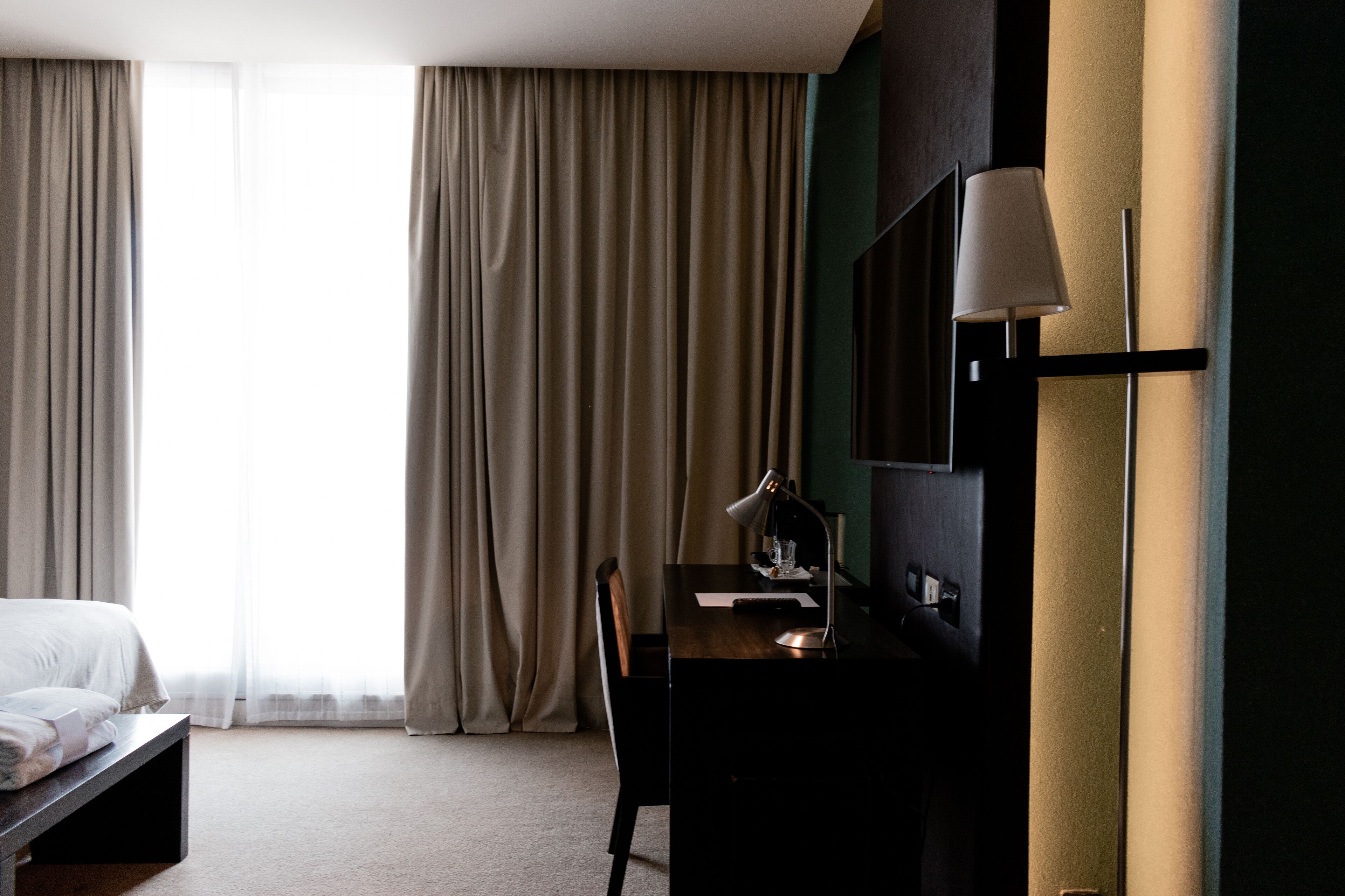 A hotel room with a bed, a desk, a chair, a lamp, and a large window with curtains.