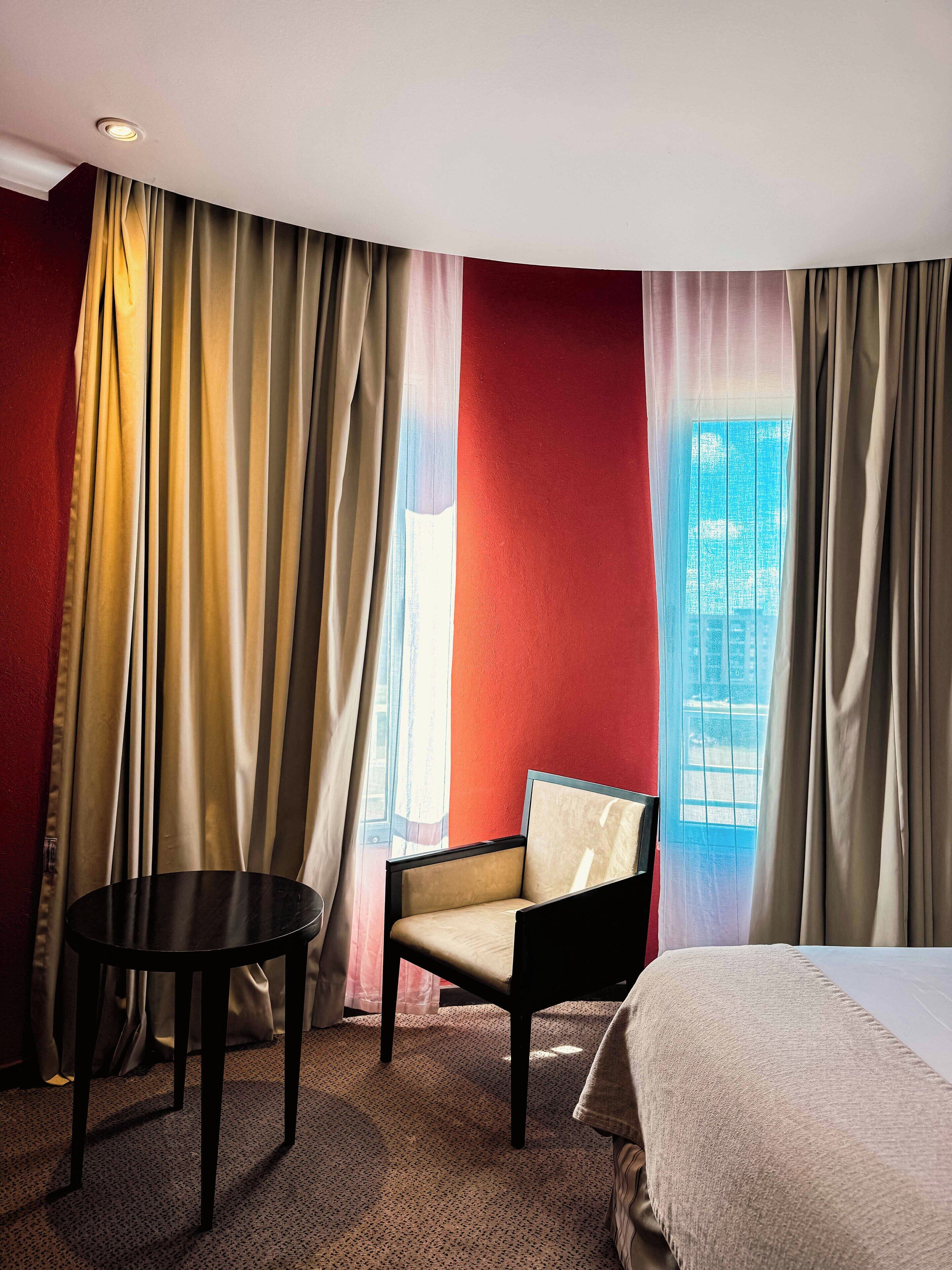 A hotel room with a bed, a chair, a small table, and a window with curtains.