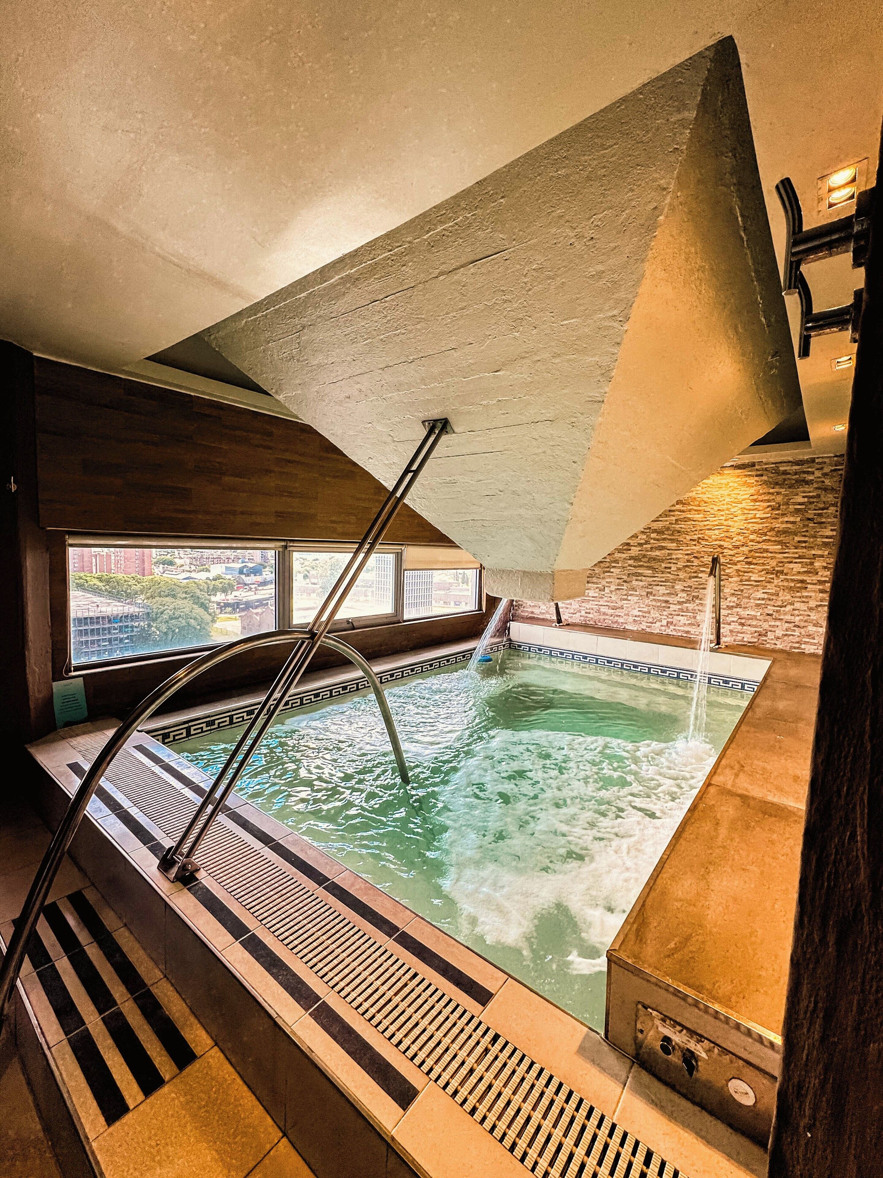 Una piscina interior con techo inclinado, una escalera que baja al agua y vistas al exterior a través de amplios ventanales.