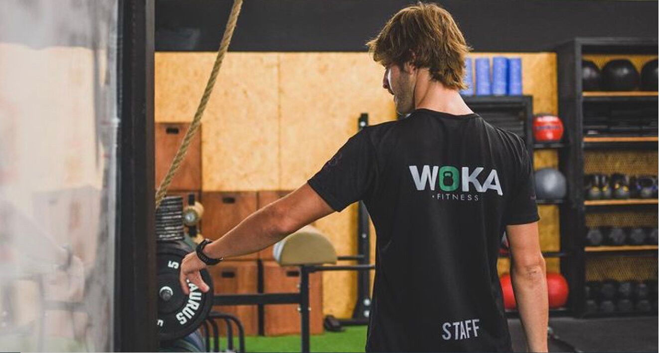 Un gimnasio con una persona usando una camiseta de 'WOKA Fitness', equipo de levantamiento de pesas y pelotas de ejercicio al fondo.