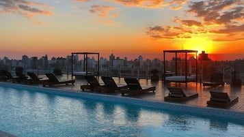 A rooftop pool with lounge chairs and a city skyline at sunset.