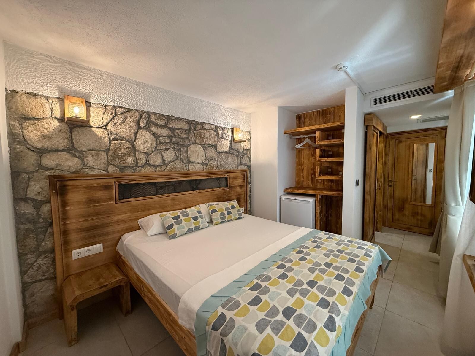 A bedroom with a stone wall, wooden headboard, bedside table, bed with patterned bedding, built-in wooden shelving, and a small refrigerator.
