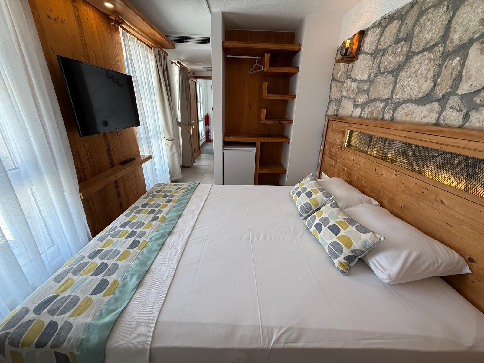A hotel room with a bed, a TV, wooden shelves, and a stone wall.