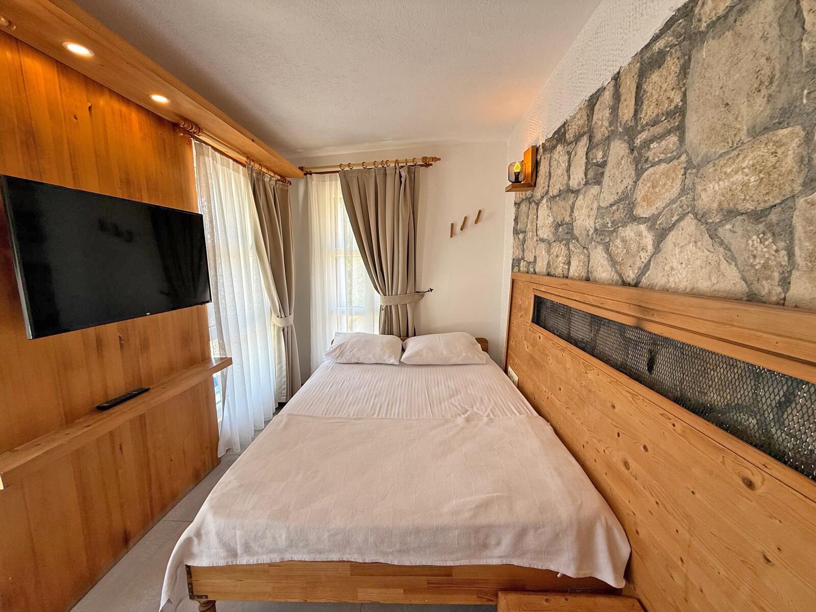 A bedroom with a bed, wooden headboard, stone wall, and a TV mounted on the wall.