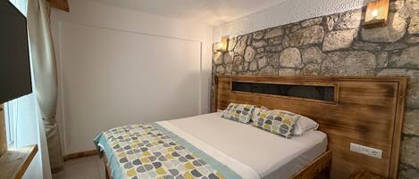 A bedroom with a stone wall, a wooden headboard, a bed with patterned bedding, a bedside table, and a small wooden stool.