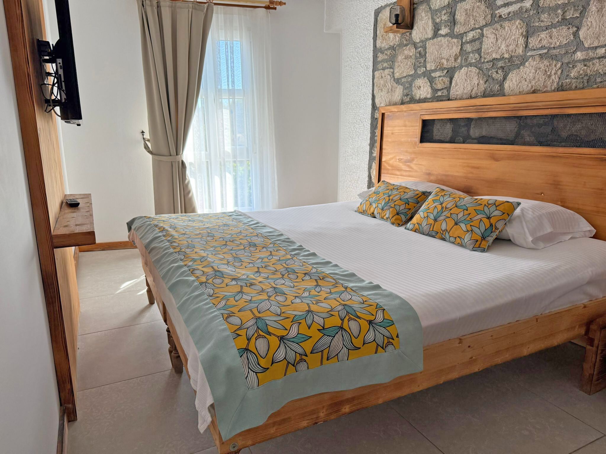 Quarto com cama de madeira, parede de pedra, janela com cortinas e televisão fixada na parede.