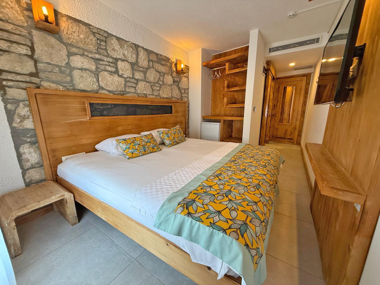 A bedroom with a stone wall, wooden headboard, a bed with patterned bedding, a bedside table, and a wooden wardrobe.