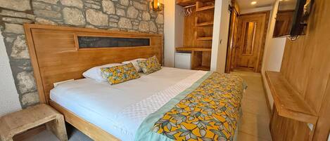 A bedroom with a stone wall, wooden headboard, a bed with patterned bedding, a bedside table, and a wooden wardrobe.