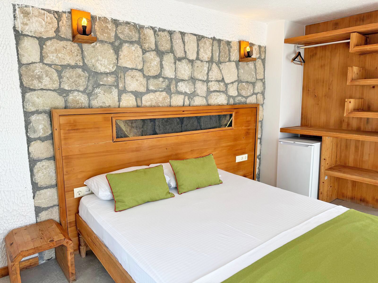 A bedroom with a stone wall, wooden headboard, two green pillows, a white bedspread, and a small wooden stool.
