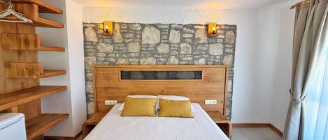 A bedroom with a stone wall, wooden headboard, two bedside lamps, a bed with white bedding and two yellow pillows, and a wooden shelf unit.