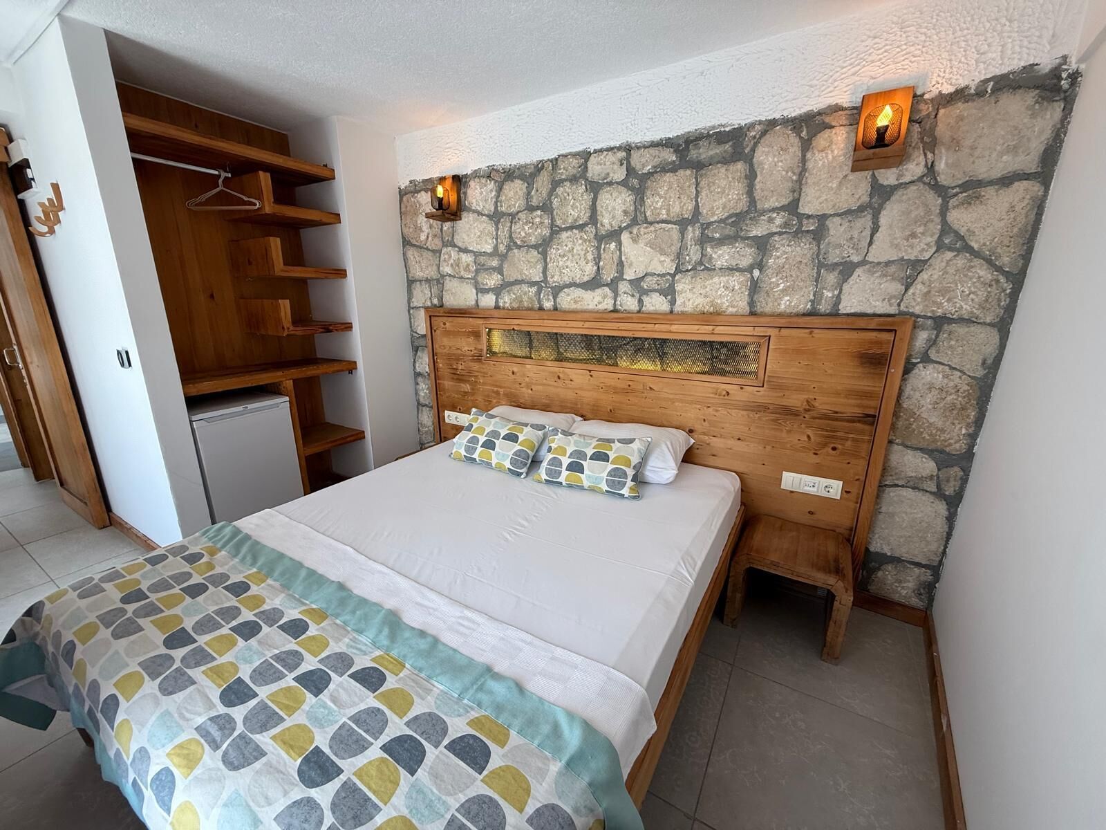 A bedroom with a bed, a stone wall, a wooden headboard, a nightstand, and a wardrobe with shelves.