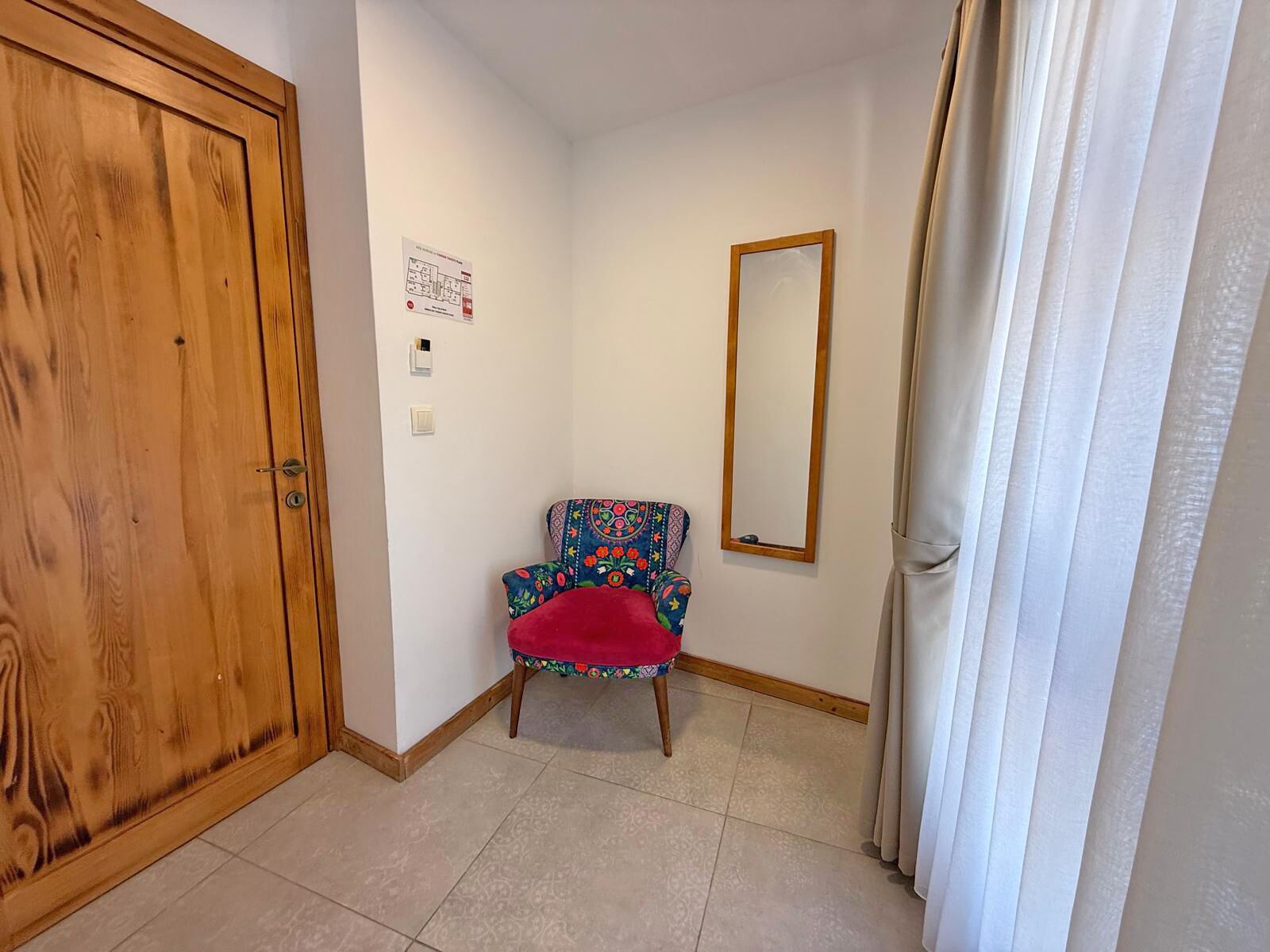 A room with a wooden door, a chair with a colorful cushion, a mirror on the wall, and a white curtain.