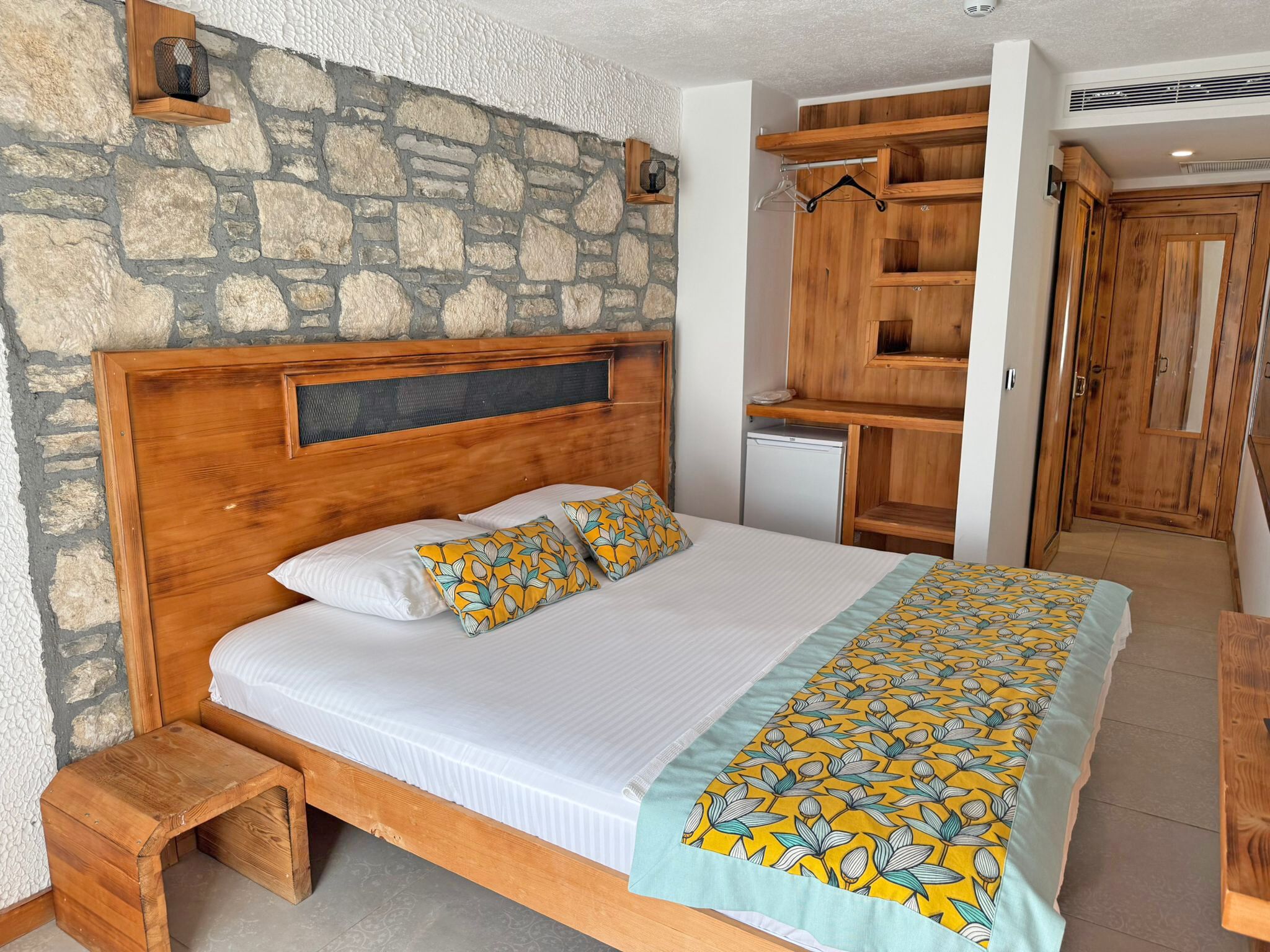 A bedroom with a stone wall, wooden bed frame, white bedding, a small wooden stool, and a built-in wardrobe.