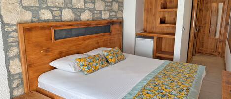 A bedroom with a stone wall, wooden bed frame, white bedding, a small wooden stool, and a built-in wardrobe.