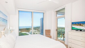 Ein Schlafzimmer mit einem gro ßen Bett, einem Fernseher, einem Sessel und Blick auf das Meer und Gebäude.