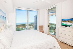Une chambre avec un grand lit, une télévision, un fauteuil et une vue sur l’océan et les bâtiments.